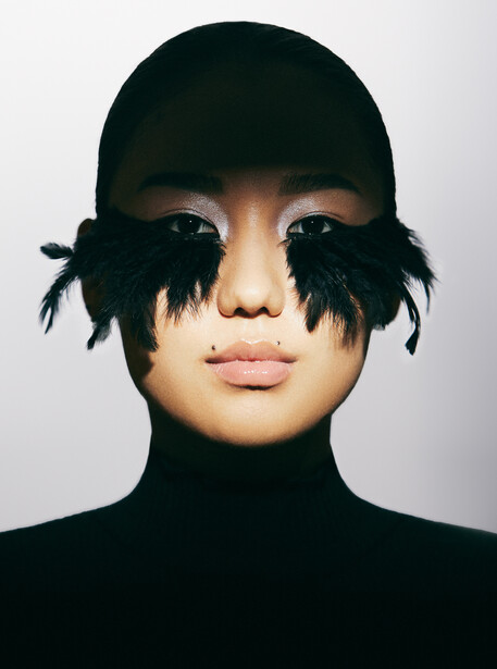 Person wearing black cap and turtleneck with dramatic black feather eye covering against neutral background.