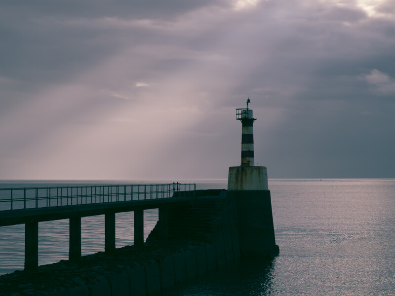 Silhouetted lighthouse and pier structure extending into calm sea under dramatic cloudy sky.