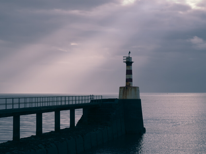 Striped lighthouse tower at the end of a concrete pier extending into calm sea under overcast sky.