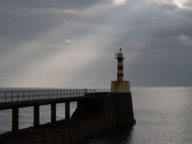 Striped lighthouse on a jetty extending into calm water under dramatic overcast sky.