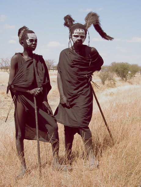 Two people with body paint standing in dry grassland, holding walking sticks and looking toward camera.