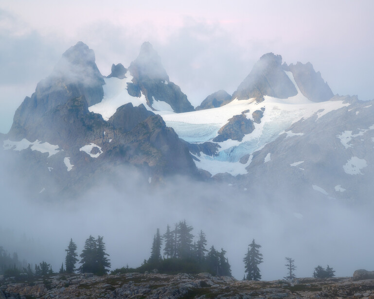 Snow-covered mountain peaks rising through dense fog and mist, with forested slopes below.