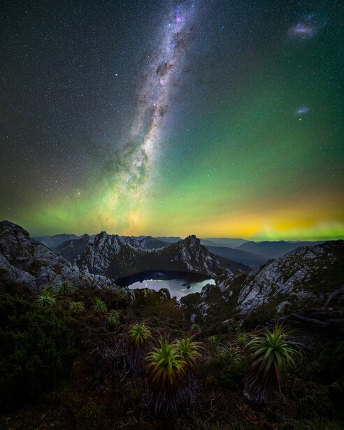 Milky Way arcing across night sky above mountains and lake with aurora borealis glow.