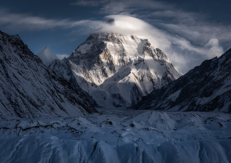 Snow-covered mountain peak framed by glaciated valleys under dramatic cloudy sky.