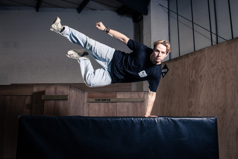 Man performing a horizontal kick mid-air above a blue padded surface in an industrial studio space.