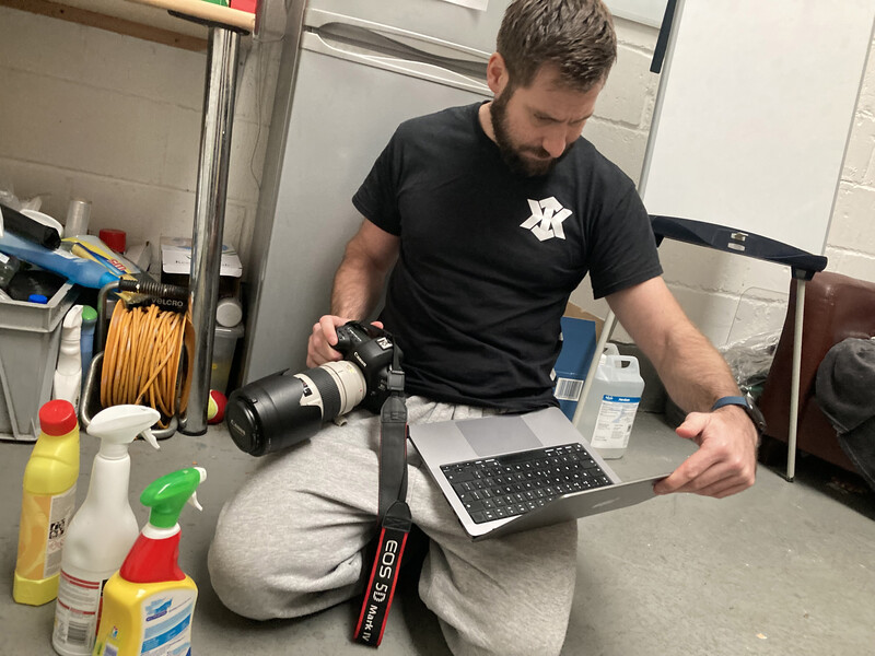 Photographer working at laptop while holding a camera lens, surrounded by production gear and supplies.