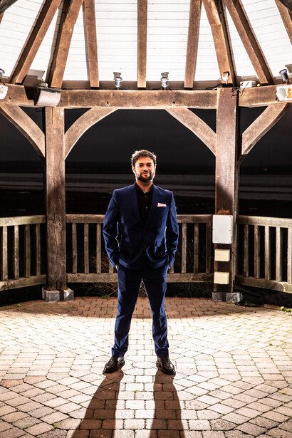 Man in navy athletic jacket standing in geometric shadow on brick courtyard beneath wooden pavilion structure.