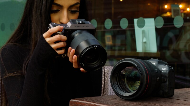 Female photographer holding a mirrorless camera with telephoto lens attached, with second camera body on table.