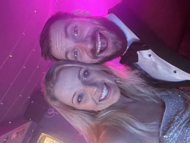 Two people smiling at camera in formal attire under magenta ambient lighting.