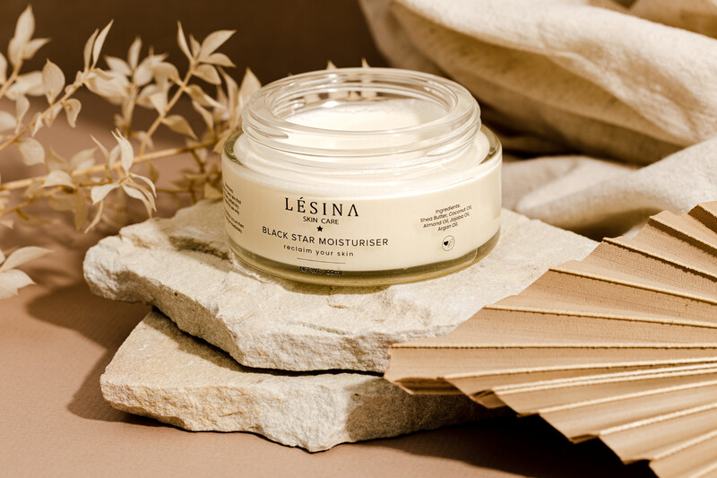 Cream-colored moisturizer jar on white stone with dried botanicals and woven elements.