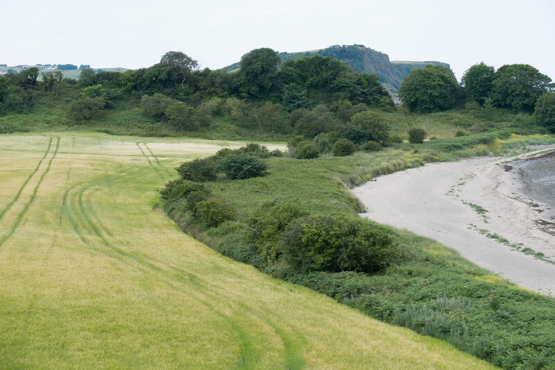 Curved crop lines in a green field beside a meandering river with vegetation and distant hills.
