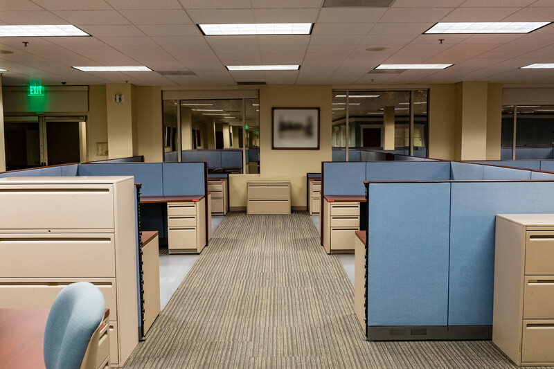 Empty office cubicles with blue dividers and beige filing cabinets in a fluorescent-lit workspace.