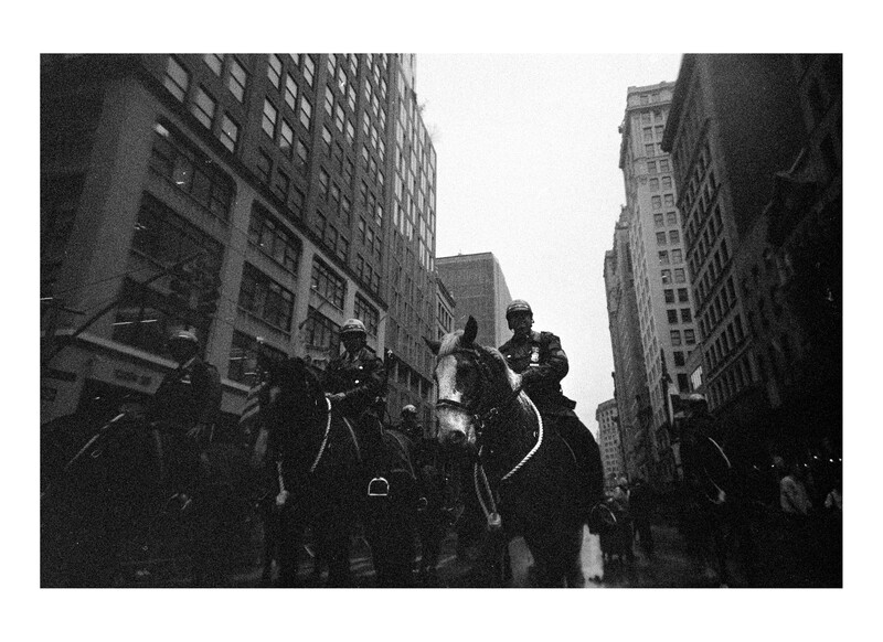 Mounted police officers riding horses down a narrow urban street lined with tall buildings.