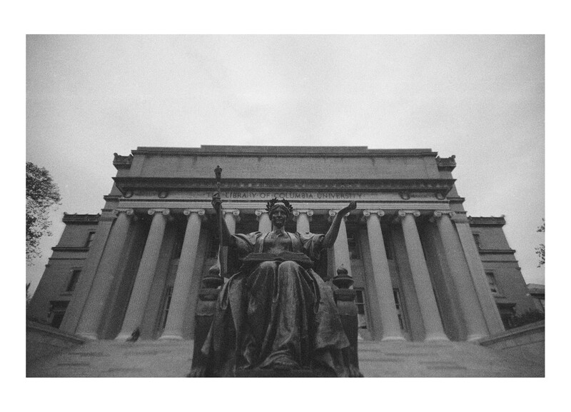 Statue of a robed female figure with outstretched arms before a neoclassical building with tall columns.