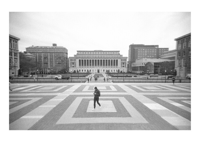 Person walking across a geometric striped plaza toward classical civic buildings in black and white.