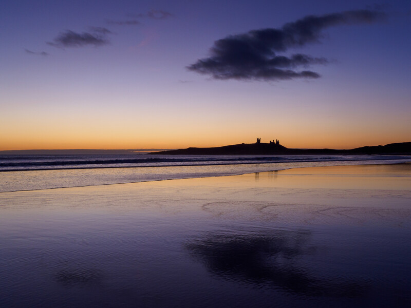Silhouetted castle ruins on a distant headland at twilight, reflected in wet sand beach.
