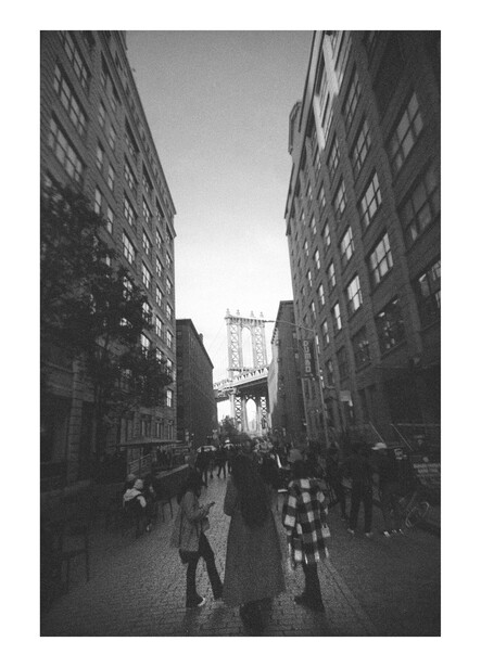 Urban street canyon with tall brick buildings framing a distant bridge view, pedestrians walking on wet pavement.