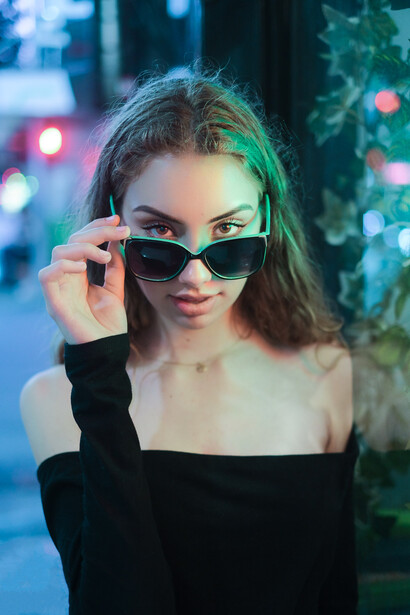 Woman wearing cat-eye sunglasses and off-shoulder black top against neon-lit urban backdrop.