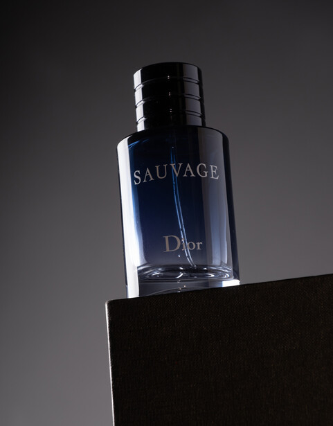 Blue glass fragrance bottle with black cap positioned on black cube against gray background.