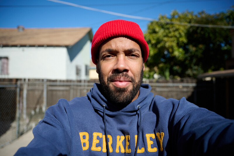 Man wearing red beanie and blue hoodie smiling at camera in bright daylight.