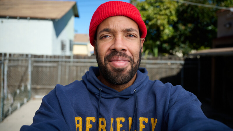 Man wearing red beanie and navy Berkeley hoodie smiles at camera in outdoor residential setting.