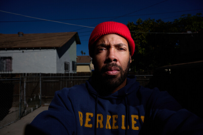 Man wearing a red beanie and dark Berkeley hoodie, photographed outdoors during golden hour with a residential building and fence visible behind him.