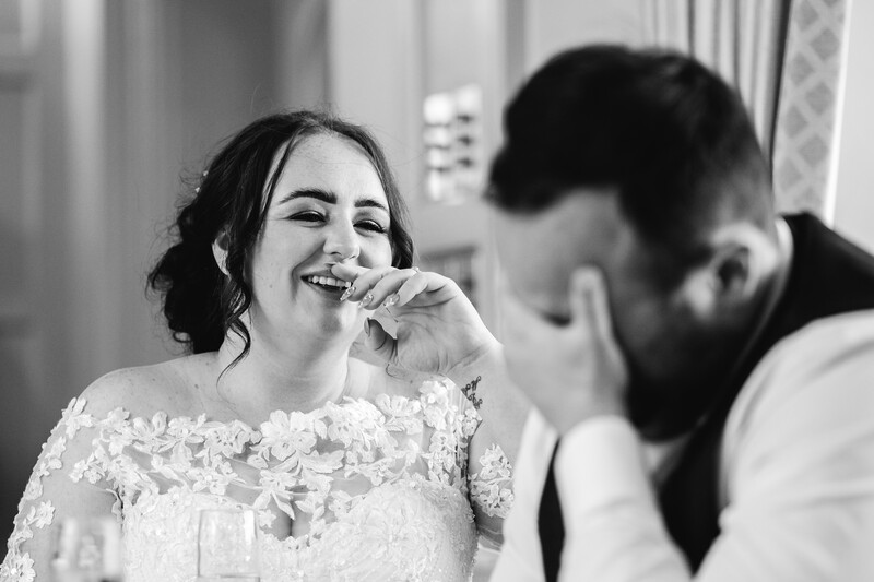 Woman in lace white dress laughing while eating, with man in dark shirt beside her at an indoor event.