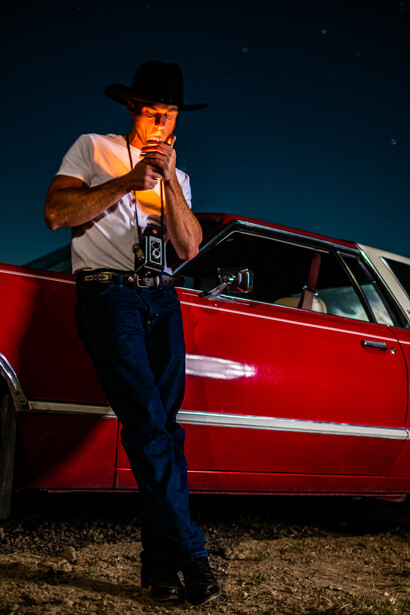 Man in cowboy hat and white shirt leaning against red vintage car at night, holding a camera to his face.