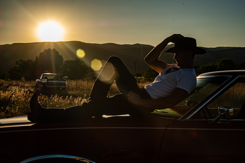 Silhouetted person relaxing in convertible car during golden hour with mountains and sun in background.