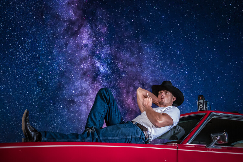 Man in cowboy hat lying on red car roof against starry night sky with Milky Way.