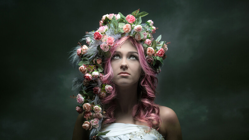 Woman with pink hair wearing elaborate floral crown against dark background.