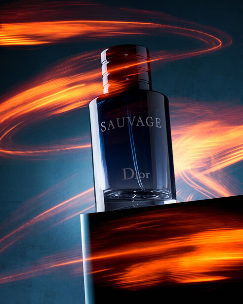 Dark fragrance bottle with white cap positioned against dynamic orange and red light streaks on dark blue background.