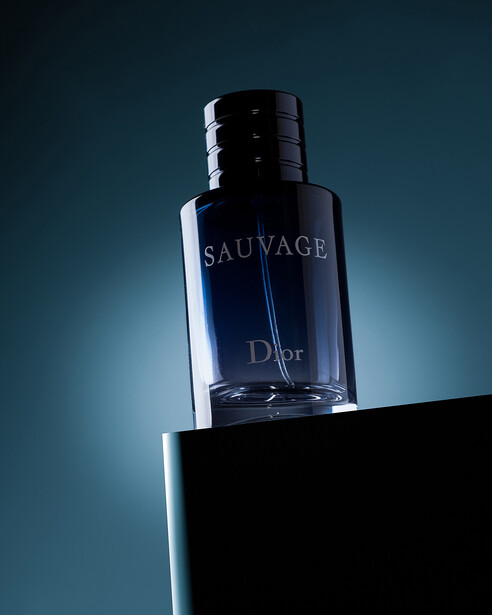 Dark glass fragrance bottle with black cap positioned on a geometric black platform against teal gradient background.