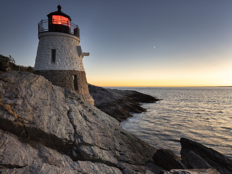 Coastal lighthouse with red beacon perched on rocky cliff at dusk.