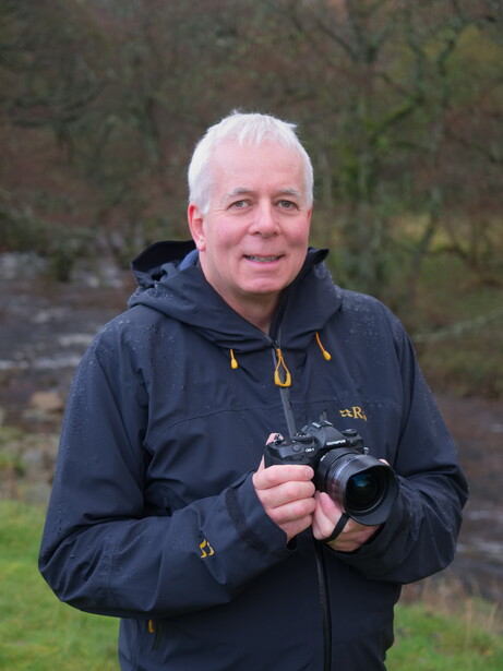 Older man holding a camera with telephoto lens in an outdoor setting with blurred green foliage background.