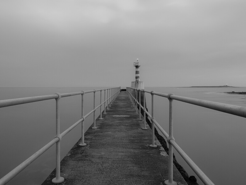 Long concrete pier with metal railings extending into calm, misty water under overcast sky.
