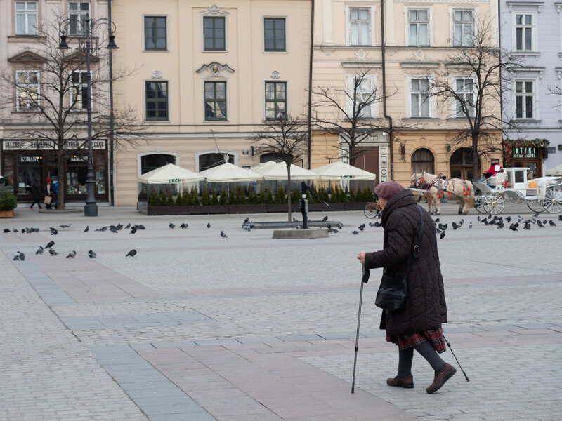 Elderly person with walking stick crossing a plaza with pigeons and historic European buildings in background.