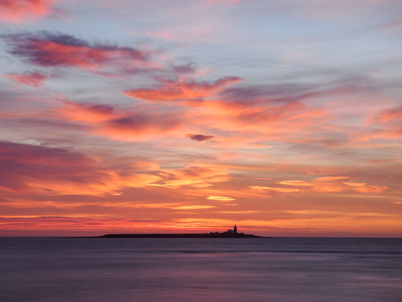 Vibrant sunset sky with orange and pink clouds over calm water, small silhouetted lighthouse on distant horizon.