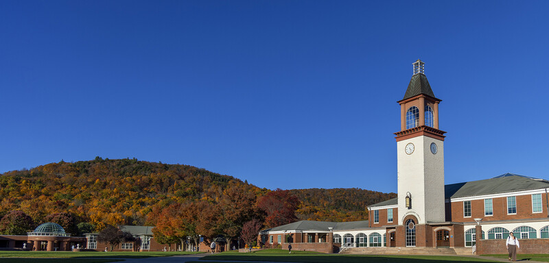 Brick academic building with tall clock tower beside autumn-colored hillside under clear blue sky.