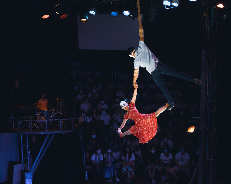 Aerial silk performer suspended in red fabric against dark venue with stage lighting.