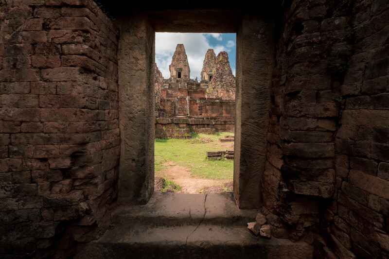 Ancient temple ruins viewed through a stone doorway frame, with towers visible across a grassy courtyard.