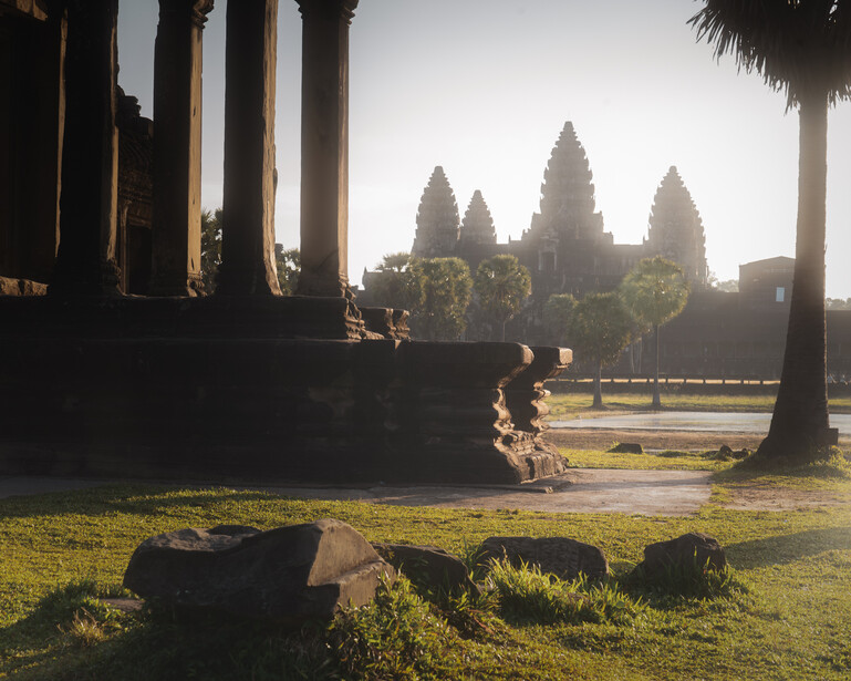 Ancient temple with four tall spires viewed through columns and palm trees at golden hour.