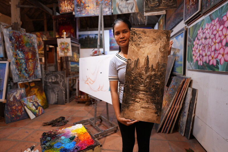 Young artist holding a textured gold-toned painting in a studio surrounded by colorful artworks.