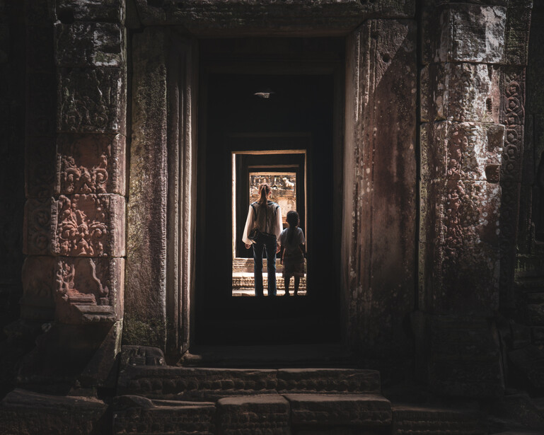 Couple standing in a doorway framed by weathered stone archway, backlit by warm light from outside.
