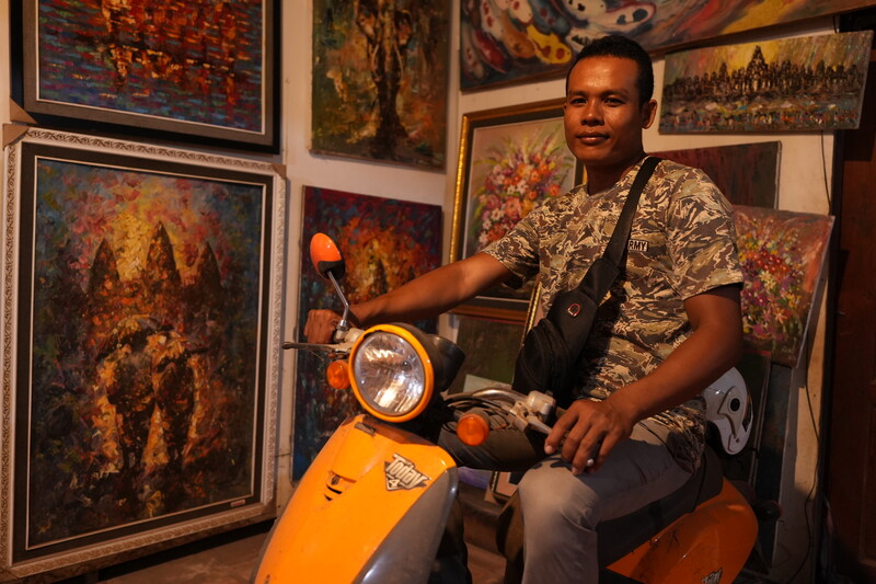 Man sitting on an orange scooter posing in front of gallery walls displaying large colorful paintings.
