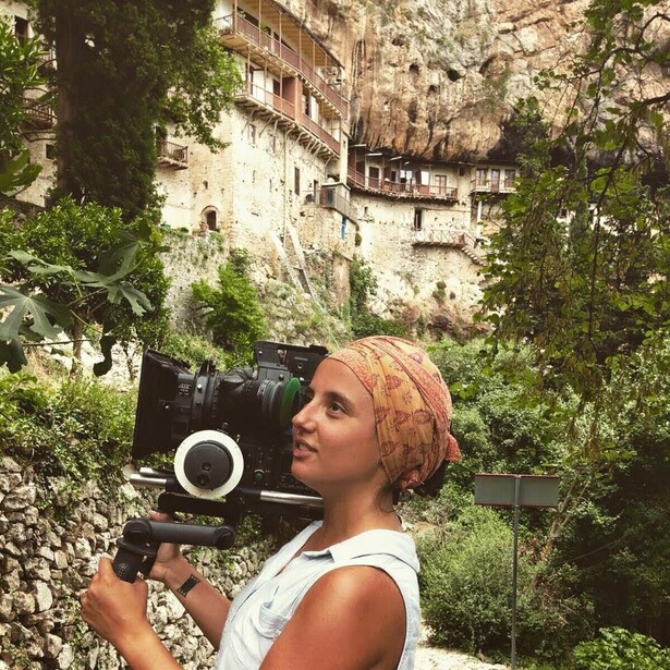 Filmmaker holding a professional cinema camera against her shoulder, photographed outdoors with historic stone architecture in the background.
