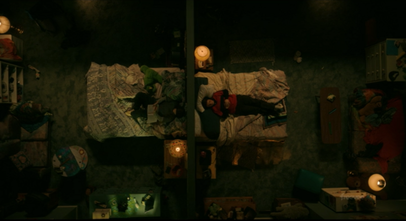 Overhead view of a dimly lit interior space with figures, fabrics, and warm candlelight creating dramatic shadows.