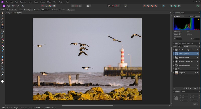 Flock of geese in flight over yellow rocks with a lighthouse in soft focus in the background.