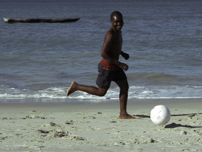 Young athlete kicking a soccer ball on a sandy beach with ocean waves in the background.