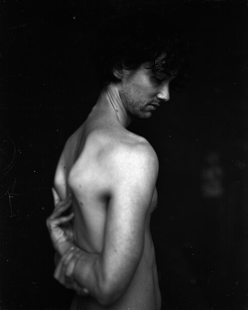 Shirtless man photographed from behind against a dark background, looking over shoulder.